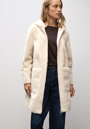 Lys beige faux fur frakke med hætte, der har to forlommer og en frontlynt. Båret over en mørkebrun top og blå jeans.