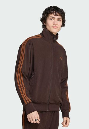 Joven con pelo rizado que lleva una chaqueta deportiva marrón con cremallera y rayas naranjas en las mangas, y pantalones a juego, sobre un fondo liso.
