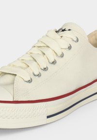 Κοντινό ενός λευκού low-top καμβά παπουτσιού με λευκά κορδόνια, μεταλλικά ακροκέρατα, κόκκινες και μπλε ρίγες στη σόλα και λαστιχένια μύτη.