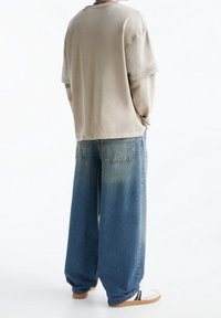 Personne portant une chemise beige à manches longues superposée, un jean bleu délavé à jambes larges et des baskets blanches à semelles en caoutchouc, debout les mains dans les poches.