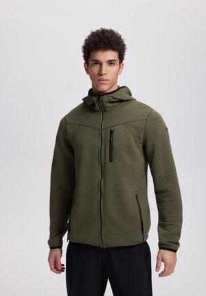 Mies pukeutuneena oliivinvihreään vetoketjulliseen hupulliseen takkiin, jossa on mustat vetoketjulliset taskut. Hän seisoo yksinkertaisen vaaleanharmaan taustan edessä.