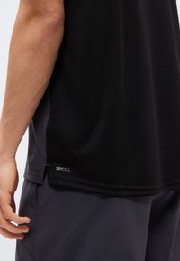 Κοντινό πλάνο ενός ατόμου που φοράει μαύρο πουκάμισο με την επιγραφή "DRYCELL" κοντά στο τελείωμα και σκουρόχρωμο σορτς, που δείχνει το αριστερό χέρι και το χέρι χαλαρά δίπλα στο σώμα.