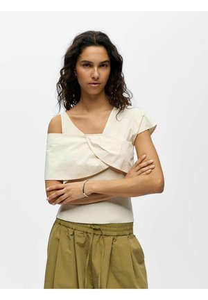 Femme aux cheveux bouclés et foncés porte un haut asymétrique blanc cassé sur un débardeur côtelé et un pantalon ample vert olive avec un cordon.
