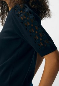 Gros plan sur un haut noir avec des motifs géométriques découpés sur la manche courte, porté par une personne aux cheveux bouclés.