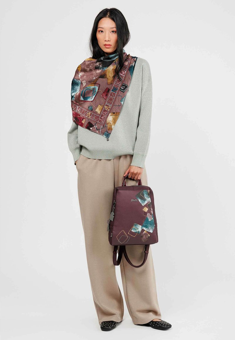 Mochila color burdeos con diseños abstractos en verde azulado y beige; material de cuero texturizado; combinada con un suéter gris claro y una bufanda estampada.