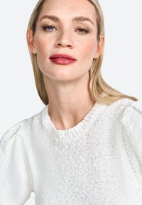 Bílý pletený svetr s texturovaným povrchem a žebrovaným výstřihem. Má nabírané rukávy a volný střih. Modelka má neutrální líčení.