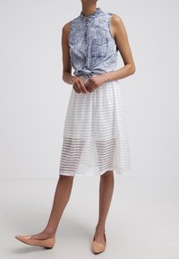 Femme portant une chemise sans manches délavée à boutons nouée à la taille, une jupe midi blanche transparente à rayures, et des ballerines pointues couleur peau, debout devant un fond blanc.