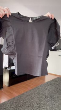 Zwarte blouse met korte mouwen en ruches, vastgehouden door twee handen in een woonkamer met een houten vloer en een grijze vloerkleed.