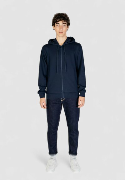 Felpa con zip di colore blu navy, con lacci, maniche lunghe, polsini e orlo a costine; indossata sopra jeans scuri e sneakers bianche. Texture semplice e liscia.