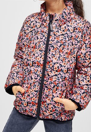 Niño vistiendo una chaqueta acolchada floral colorida, con las manos en los bolsillos, abotonada, combinada con jeans de mezclilla oscuros.