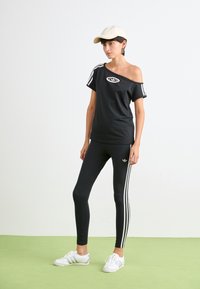 Žena stojaca na zelenej podlahe, oblečená v čiernom off-shoulder top a značky Adidas, čiernych legínach s bielymi pruhmi, bielych teniskách a béžovej čiapke.