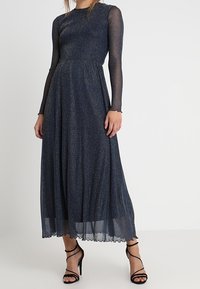 Robe longue marine en tissu pailleté, avec de longues manches transparentes, un corsage ajusté et une jupe fluide à l'ourlet en forme de vagues. Sandales à talons noires.