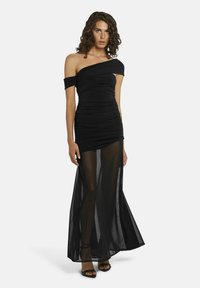 Vrouw in een zwarte one-shoulder ruched jurk met een doorschijnende lange rok, staand en met zwarte hoge hakken.