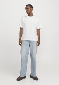 Witte katoenen T-shirt met korte mouwen, ronde hals en een klein logo-detail. Gecombineerd met lichtblauwe versleten jeans en donkerbruine schoenen.