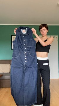 Denim brezrokavnik v temno modri barvi; ima gumbe spredaj, žep na prsih in dolg, ohlapen kroj. Tkanina je postavljena navzgor za prikaz.