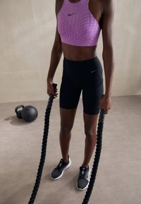 Shorts de sport noirs et brassière de sport violette avec un motif géométrique, tenant des cordes de combat noires. Chaussures de sport noires avec semelles blanches.