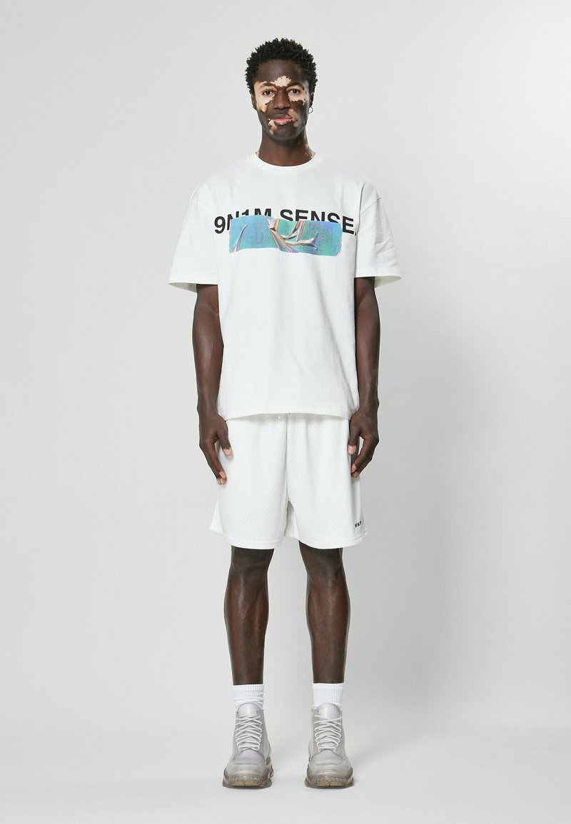 T-shirt en coton blanc avec impression graphique et texte noir, associé à un short en maille blanc. Les chaussures sont de couleur gris clair avec une finition texturée.