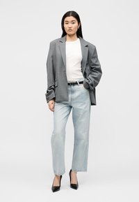 Γυναίκα με μεγάλο γκρι σακάκι, άσπρο t-shirt, ανοιχτό μπλε τζιν, μαύρη ζώνη και μαύρες γόβες, στέκεται μπροστά σε απλό λευκό φόντο.