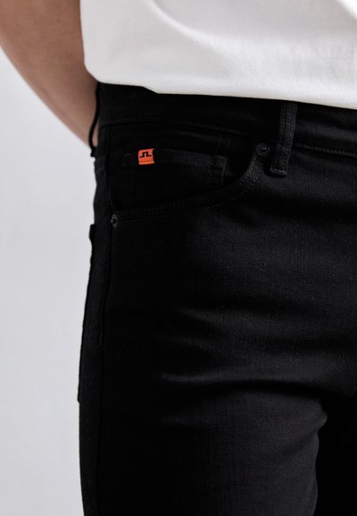 Schwarze Jeans aus Denim mit schlankem Schnitt, mit orangefarbenem Logodetail an der Vorderasche und standardmäßiger Knopfverschluss. Glatte Textur, klassisches Design.