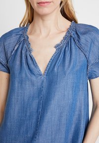 Blå denimtröja med V-ringning och draperad halsringning. Kort ärm och avkopplad passform. Mjuk textur och fransdetaljer längs kanterna.