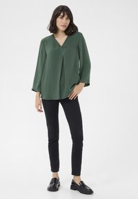 Grøn, langærmet, v-hals bluse med plisseringer, parret med sorte slim-fit bukser og blanke sorte loafers, sat imod en hvid baggrund.