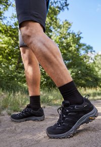 Black hiking shoes with a textured upper, lace closure, and Vibram outsole, paired with black socks and gray shorts, on a dirt path.