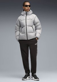 Pujsasta jakna svetlo sive barve s prešitim vzorcem, črnim zadrgo, kombinirana s črnimi trenirkami in črnimi supergami z belim podplatom.