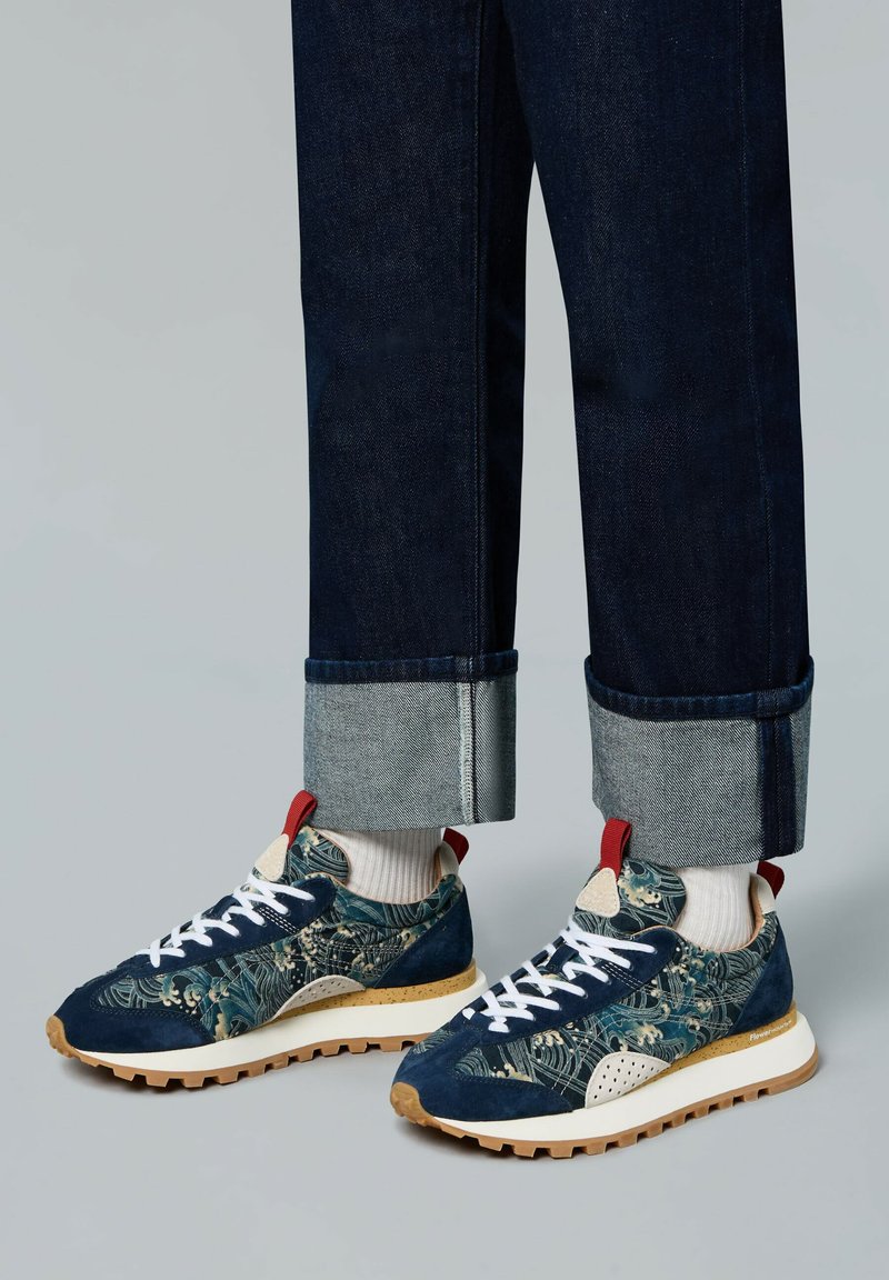 Sneakers feature a navy blue fabric with floral patterns, white laces, red heel tab, and a beige outsole. Worn with cuffed dark denim jeans.