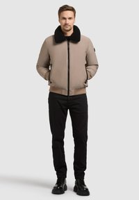 Veste bomber brun clair avec un col en fourrure noire, fermeture éclair, poches latérales et poignets élastiqués, portée sur un pantalon noir et des bottes.