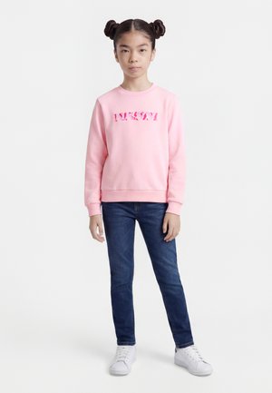 Fată tânără cu coafură cu două cocuri, purtând un hanorac roz cu text cu model, blugi albaștri și adidași albi, stând cu fața spre înainte.