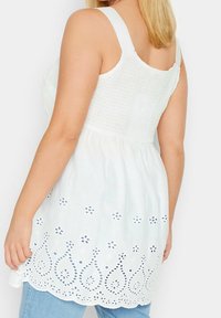 Top sans manches blanc avec un corsage smocké et un design floral brodé au niveau de l'ourlet. Tissu léger avec une silhouette fluide.