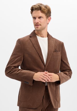 Man wearing brown corduroy suit jacket over white shirt, standing with hands clasped, looking to the side against plain background.