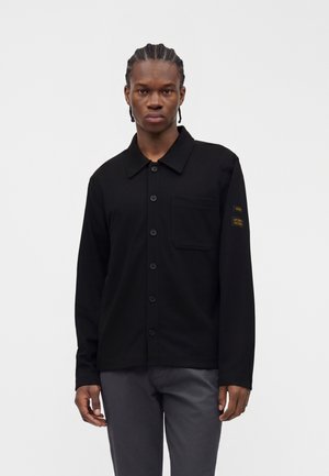 Homme aux cheveux tressés portant une chemise noire boutonnée avec une poche et un pantalon gris sur un fond uni clair.