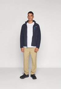 Tumesinine fliisjakk kapuutsiga, millel on sinised detailid ja lukuga taskud, kombineeritud valge t-särgi ning beežide pükstega. Mustad kingad.