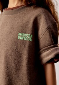 Persona che indossa una felpa marrone con texture, maniche arrotolate e testo verde "AMERICAN VINTAGE" sul petto.