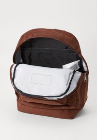 Καφέ σακίδιο από κόρντουρα με φερμουάρ στην κορυφή, ανοιχτό και αποκαλύπτοντας ένα λευκό εσωτερικό και εσωτερική τσέπη με μοτίβο εκτυπωμένου λογότυπου.