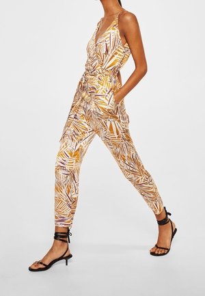 Overal s tenkými ramínky, zdobený žlutým a fialovým listovým vzorem, s úzkými nohavicemi a bočními kapsami, doplněný černými sandály s pásky.