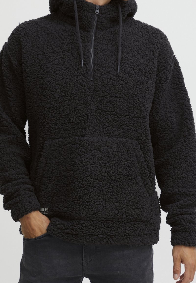 Sweat à capuche en polaire texturée noire avec poche avant et cordons, porté par une personne avec les mains partiellement visibles dans les poches.