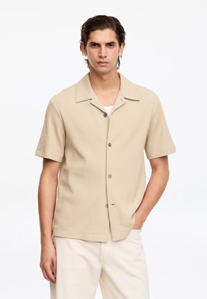 Homme portant une chemise beige texturée à manches courtes boutonnée sur un top blanc et un pantalon clair, debout avec une main dans la poche.