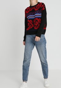 Pull noir avec des motifs floraux rouges, accents géométriques bleus, porté avec un jean bleu clair et des baskets blanches, doté d'un col rond.