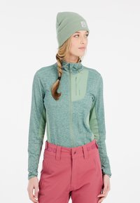 Maglione verde con zip e maniche lunghe, dalla finitura testurizzata e un pannello sul petto di un verde contrastante, abbinato a pantaloni rosa con dettagli circolari.