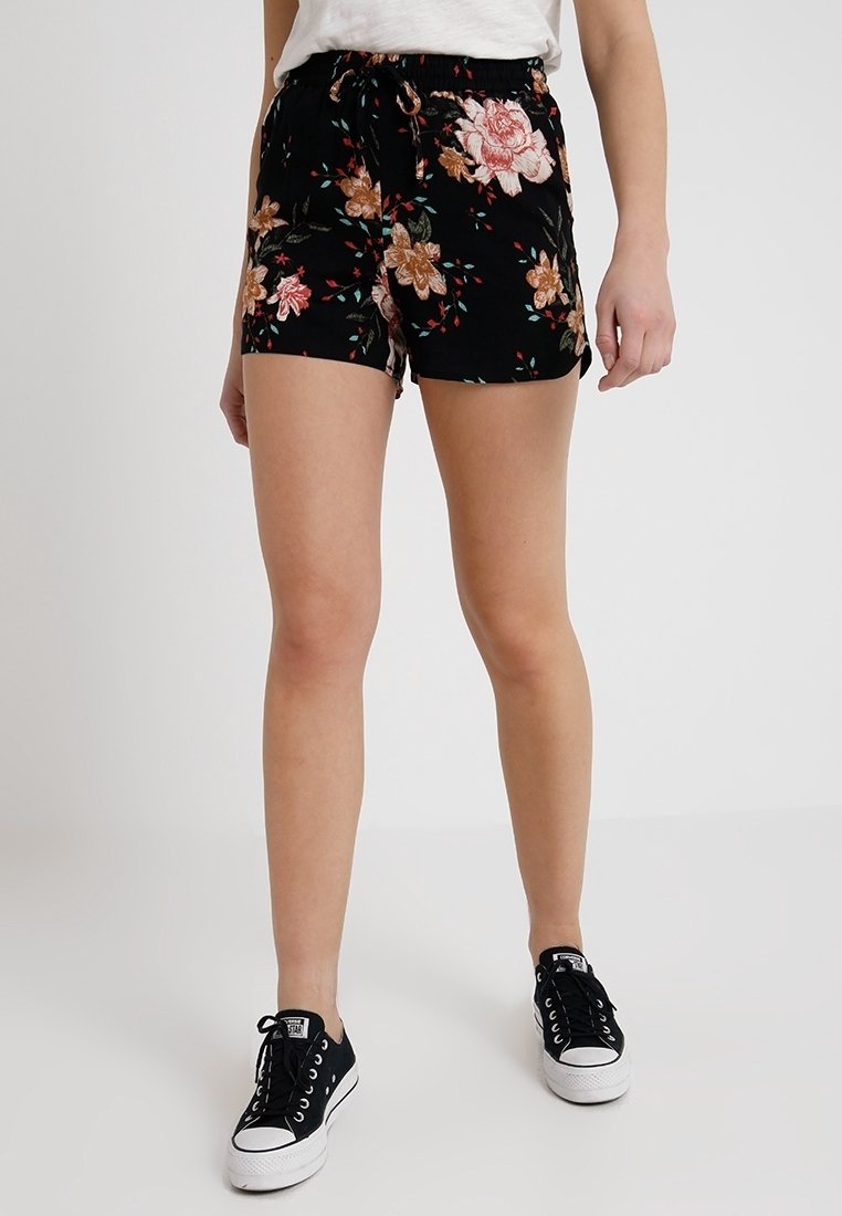 Personne portant un short noir à fleurs roses et marron, un haut blanc et des baskets basses noires, debout sur un sol blanc uni.
