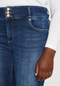 Jeans de tiro alto en azul oscuro con tres botones de cobre, bolsillo frontal visible, combinados con una camiseta de manga larga de canalé blanca y una mano descansando al costado.