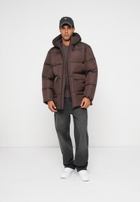 Καφέ φουσκωτό μπουφάν με κουκούλα, με ραφή καπιτονέ, δύο μπροστινές τσέπες και άνετη εφαρμογή, φορεμένο πάνω από ένα γκρι T-shirt και σκούρα τζιν.