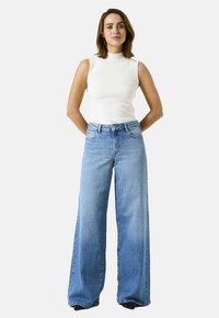 Vrouw die met haar handen achter haar rug staat, gekleed in een mouwloos wit topje en lichtblauwe wijde jeans, tegen een effen achtergrond.