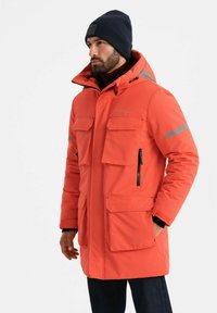 Felle oranje gewatteerde parka met meerdere voorkruiden, zwarte ritsen en reflecterende accenten. Zwarte gebreide muts op het hoofd.