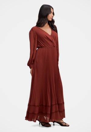 Woman with long dark hair wearing a rust-colored long-sleeve pleated maxi dress and black heeled sandals, standing and looking right.