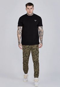 Čierne tričko s logom, maskáčové cargo nohavice v zelených tónoch a biele tenisky. Zobrazené na jednoduchom pozadí.