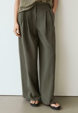 Persona che indossa pantaloni larghi verde oliva e top beige di maglia, in piedi con una mano in tasca e indossa infradito nere.