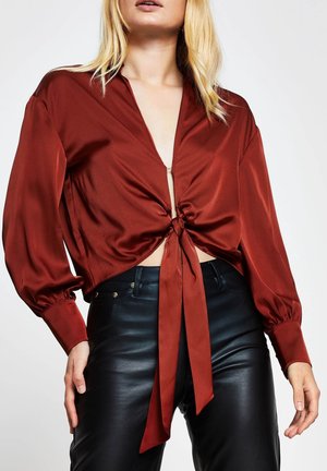Bordeauxrote Satinbluse mit tiefem V-Ausschnitt und gebundenem Vorderteil, ausgestattet mit langen, puffigenÄrmeln und einem cropped Design. Kombiniert mit schwarzen Lederhosen.